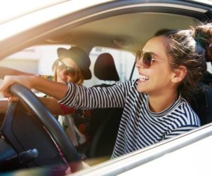 Two fun young women in sunglasses driving in a car in town laughing and smiling as they socialise together, view through open side window; Shutterstock ID 608752922; Jobnummer: -