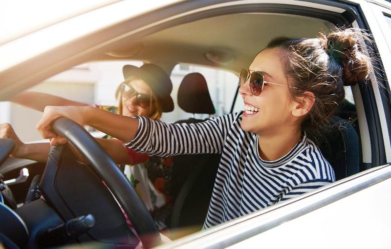 Two fun young women in sunglasses driving in a car in town laughing and smiling as they socialise together, view through open side window; Shutterstock ID 608752922; Jobnummer: -